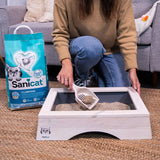 Sanicat Clumping cat litter with Oxygen Control, woman demonstrating use with scoop in wooden litter box. Packaging visible: 10L, antibacterial, dust-free.