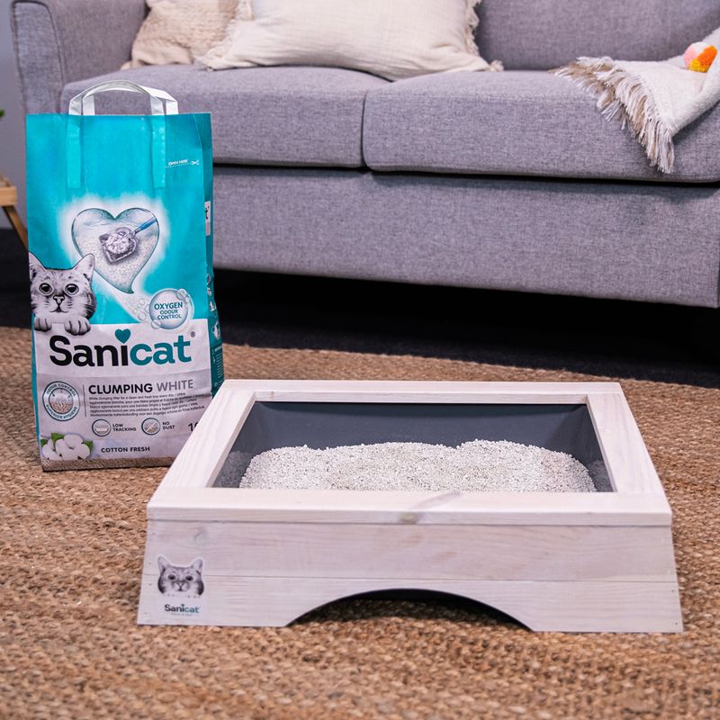 Sanicat Clumping White cat litter, Cotton Fresh scent, with oxygen control. Packaging next to a wooden litter box with litter on carpet in front of a grey sofa.