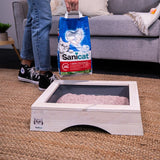 Person holding a bag of Sanicat litter above a filled litter tray, demonstrating use of the product for maintaining cleanliness.