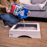 Person pouring Sanicat litter from a bag into a rectangular tray, demonstrating how to fill the tray with litter.
