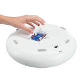 Hand opens battery compartment of a white device, three inserted AA batteries visible. Device has a round shape and grey accents.