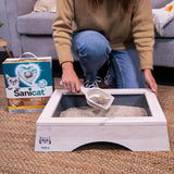 Sanicat Active Gold cat litter with oxygen technology. Woman removes clumps from white cat toilet with scoop. Litter packaging visible next to toilet.