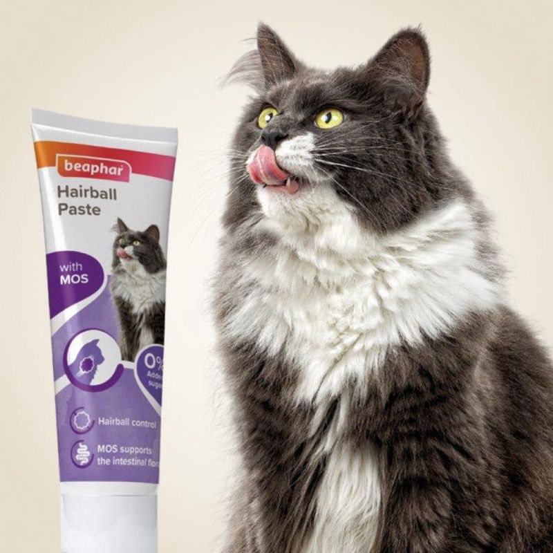 Tube labelled 'beaphar Hairball Paste with MOS', showing icons for hairball control and intestinal flora support, next to a long-haired cat licking its lips.