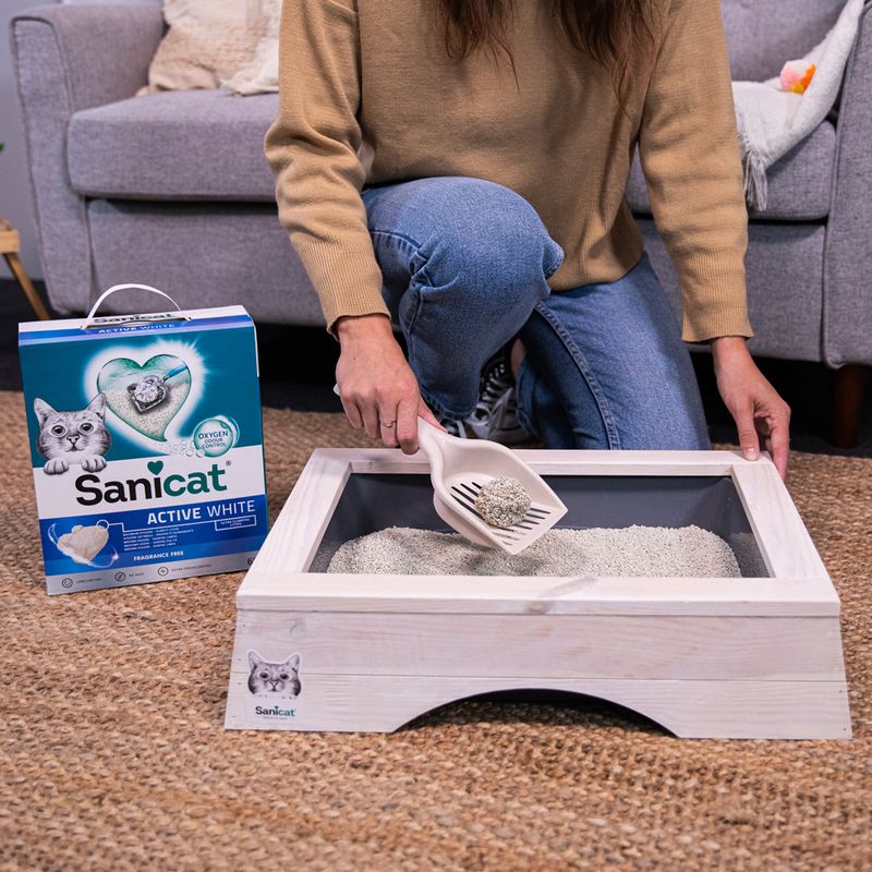Sanicat Active White cat litter, unscented. Woman demonstrates use with litter scoop in wooden cat toilet. Packaging visible with product details and cat image.