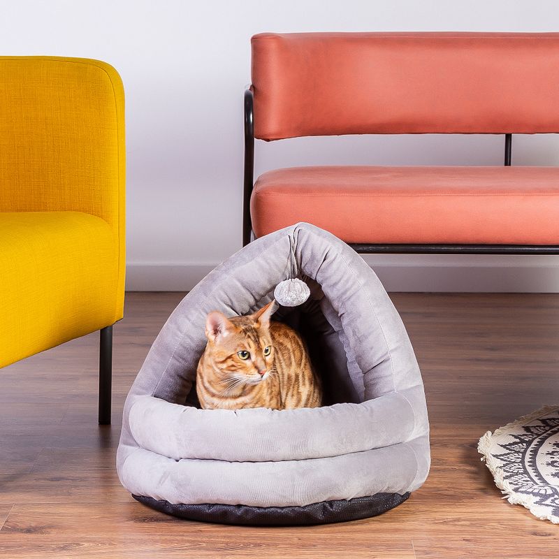 Grey, triangular cat bed with soft fabric and hanging play ball. Bengal cat sitting inside. Furniture in background: yellow armchair, orange bench.
