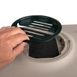 Hand lifts green lid from a round filter insert, visible is a black activated carbon filter in a beige-coloured device.