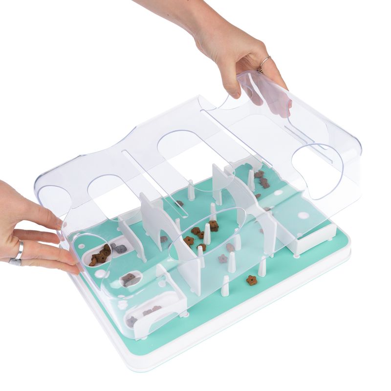 Interactive toy with transparent lid and compartments, filled with small treats. Hands open the lid for filling or removal.