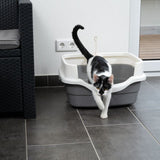 Black and white cat leaving a grey litter tray with a white rim. Litter tray on tiled floor next to a piece of furniture and a wall socket.