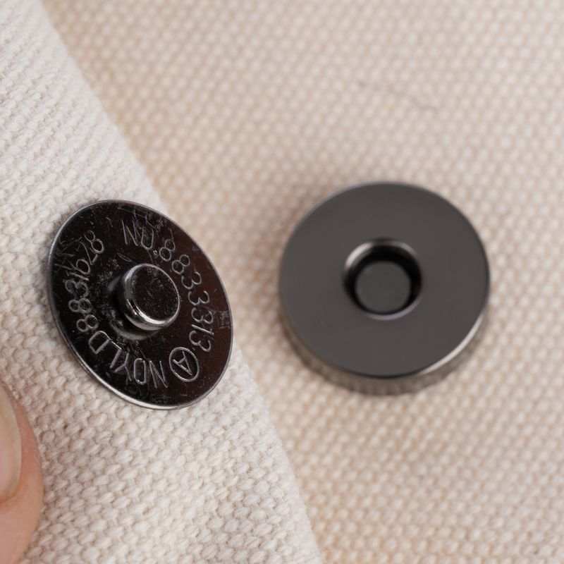 Close-up of a metal magnetic clasp on beige fabric, consisting of two parts: one with a raised centre and one flat disc.