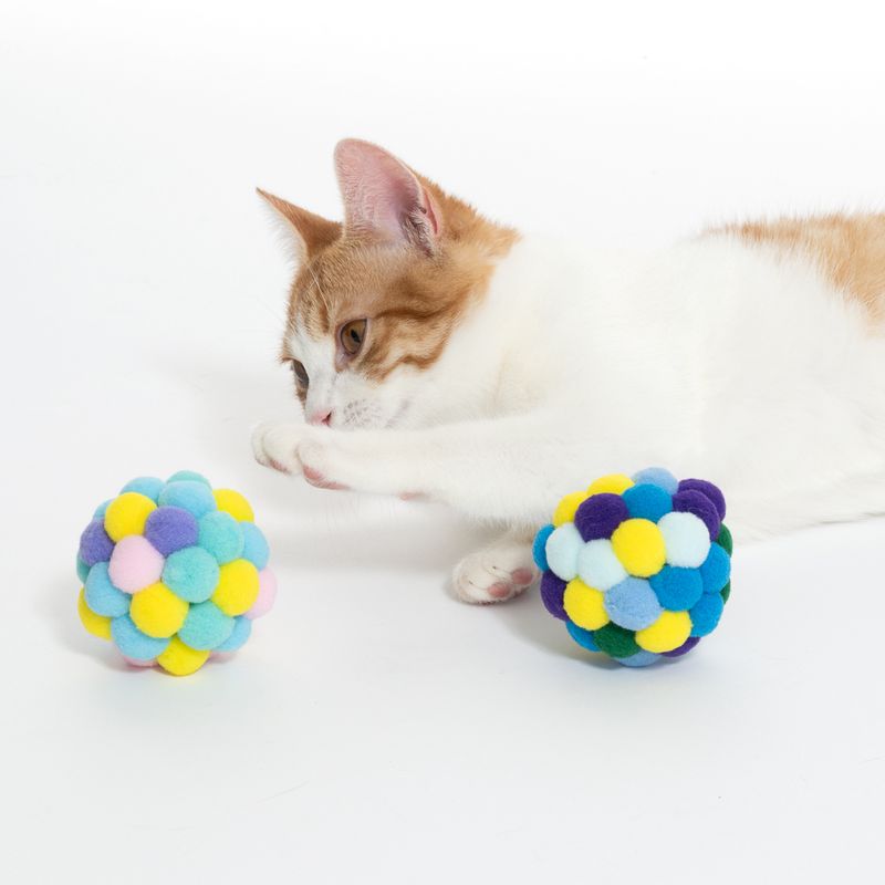 Cat playing with colourful balls made of soft fabric pom-poms. Product shows toy for cats.