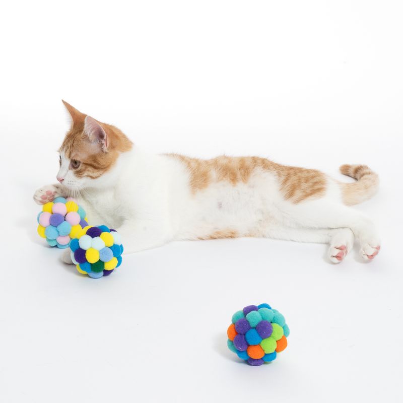 Cat playing with colourful, padded fabric balls. Three coloured play balls visible: yellow-blue, pink-purple and green-orange.