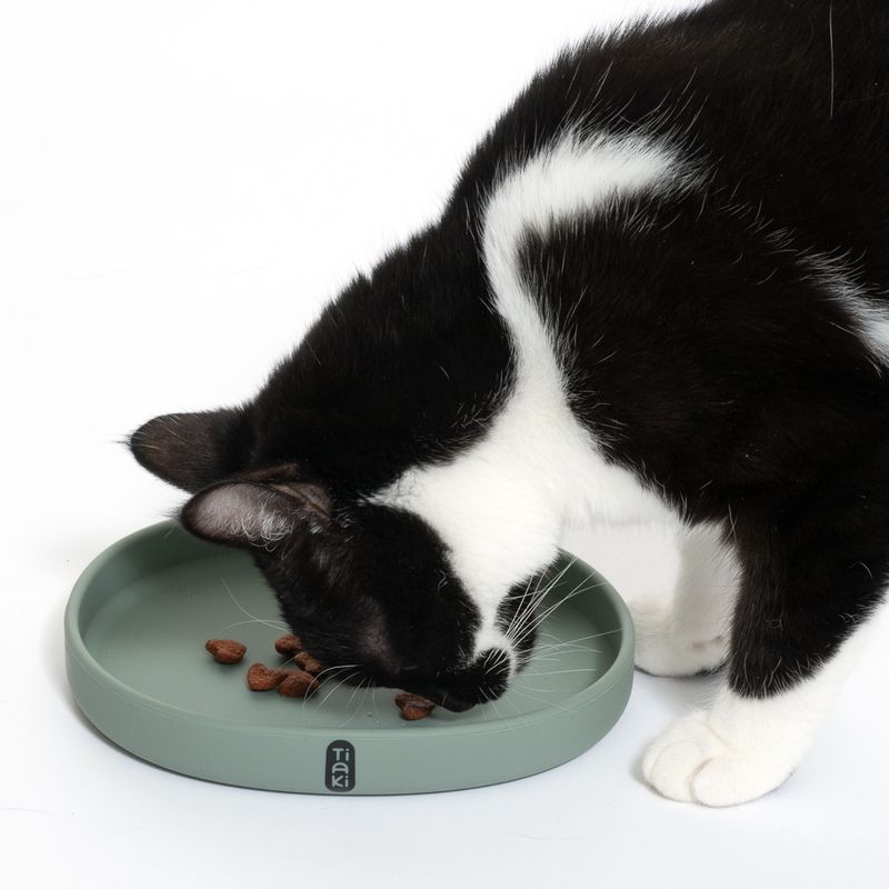 Black and white cat eating from a green food bowl with Tiaki logo. Bowl contains brown food pieces.