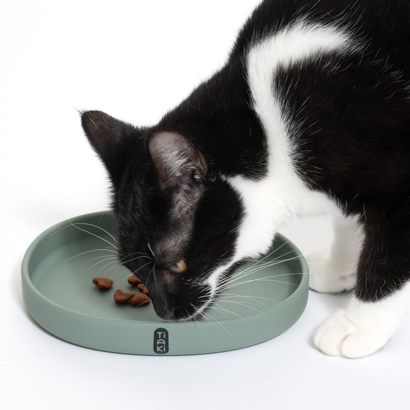 Black and white cat eating from a green food bowl with Tiaki logo. Some brown food pieces are visible in the bowl.