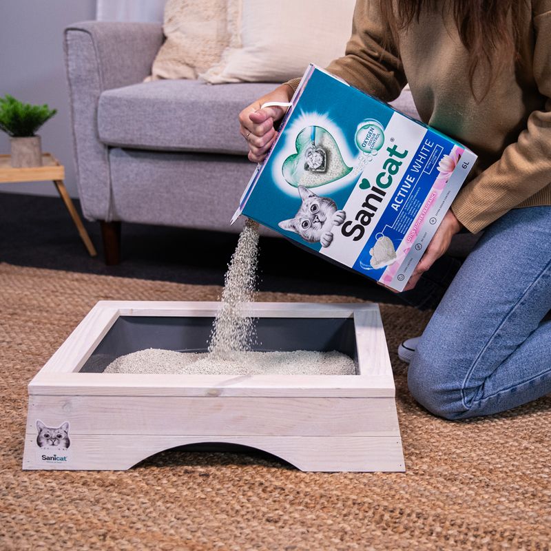 Sanicat Active White cat litter poured into a wooden cat toilet. Packaging shows a cat and heart symbol with text 'Active White'.