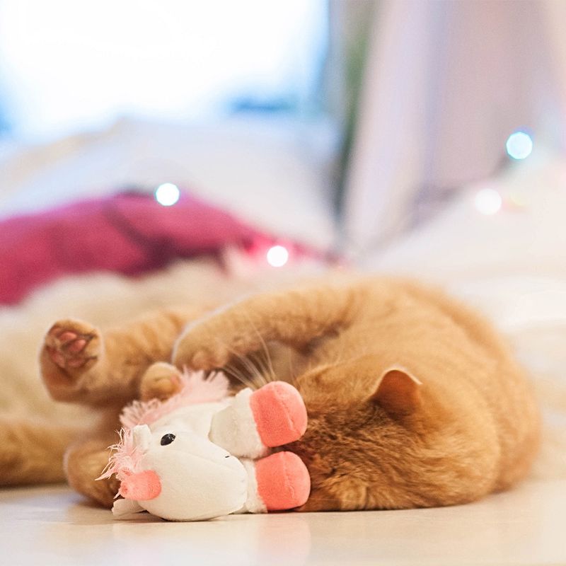 Red cat playing with white plush unicorn toy with pink accents on a light floor.