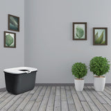 Black cat litter box with white lid and entry, placed in a room with wooden floor, green plants in white pots and wall pictures with leaf motifs.