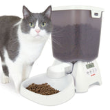 Automatic feeder with digital display, filled with dry food. White and grey cat standing next to it.