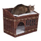 Cat basket made of brown wicker with two levels, padded top lying area and sheltered sleeping area below. Cat is sitting on the top level.