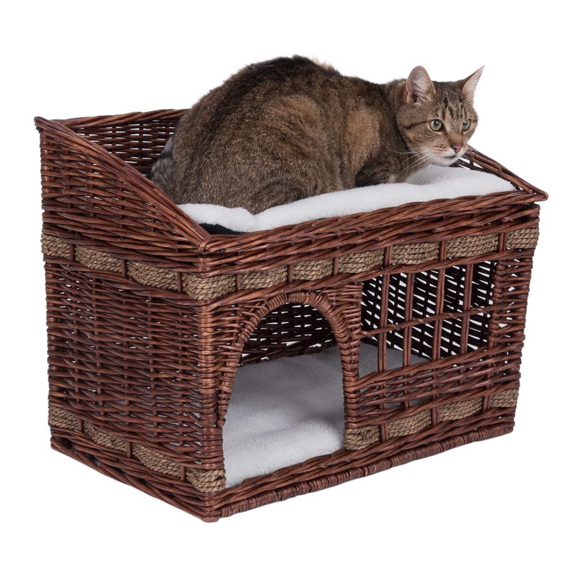 Cat basket made of brown wicker with two levels, padded top lying area and sheltered sleeping area below. Cat is sitting on the top level.