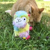 Plush toy in llama shape with colourful fur, sitting on a meadow. In the background, an orange cat in the grass.
