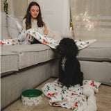 Black dog sitting on a blanket with Christmas motifs next to a green food bowl. Person holding a mug and wearing matching Christmas-themed clothing.
